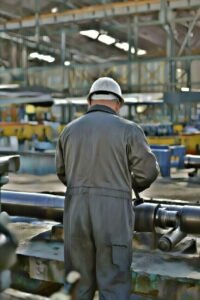 Técnico de mantenimiento industrial visto de espaldas, ajustando un rodillo largo de cinta transportadora en una fábrica con maquinaria y estructuras metálicas al fondo.